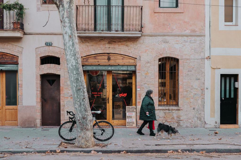 A woman walking a dog on a street in Barcelona, past the Puro Café Barceloneta. A bicycle stands by a tree, leaves on the ground. Old houses.  