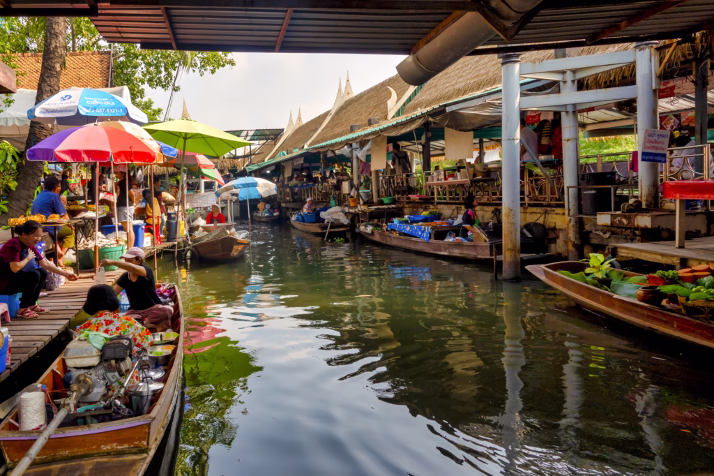 The busy Taling Chan Floating Market in Bangkok, with vendors on boats and customers buying goods at the waterside under colourful umbrellas.