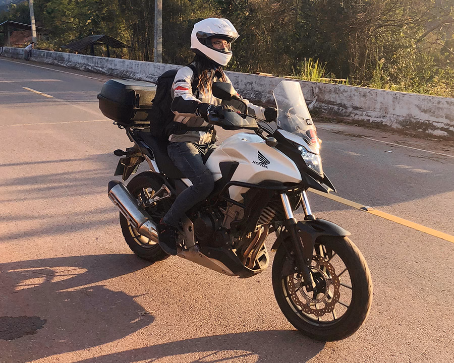 A woman in a white helmet and riding gear rides a white Honda on an asphalt road in Thailand on a sunny afternoon. Travelling by motorbike.